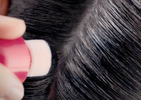 A woman holds a Garnier Express Retouch applicator uncapped and has coated half her hair with pigment while the other half remains black with streaks of gray.
